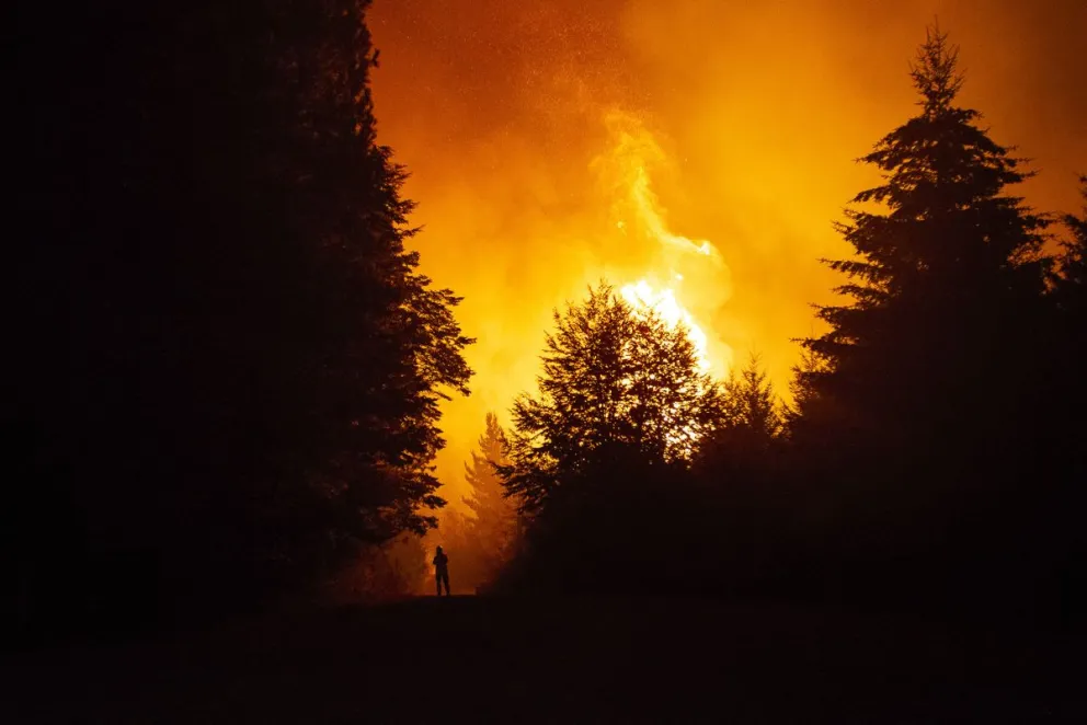 A large wildfire blazes through a forest with silhouetted trees and a person in the foreground.